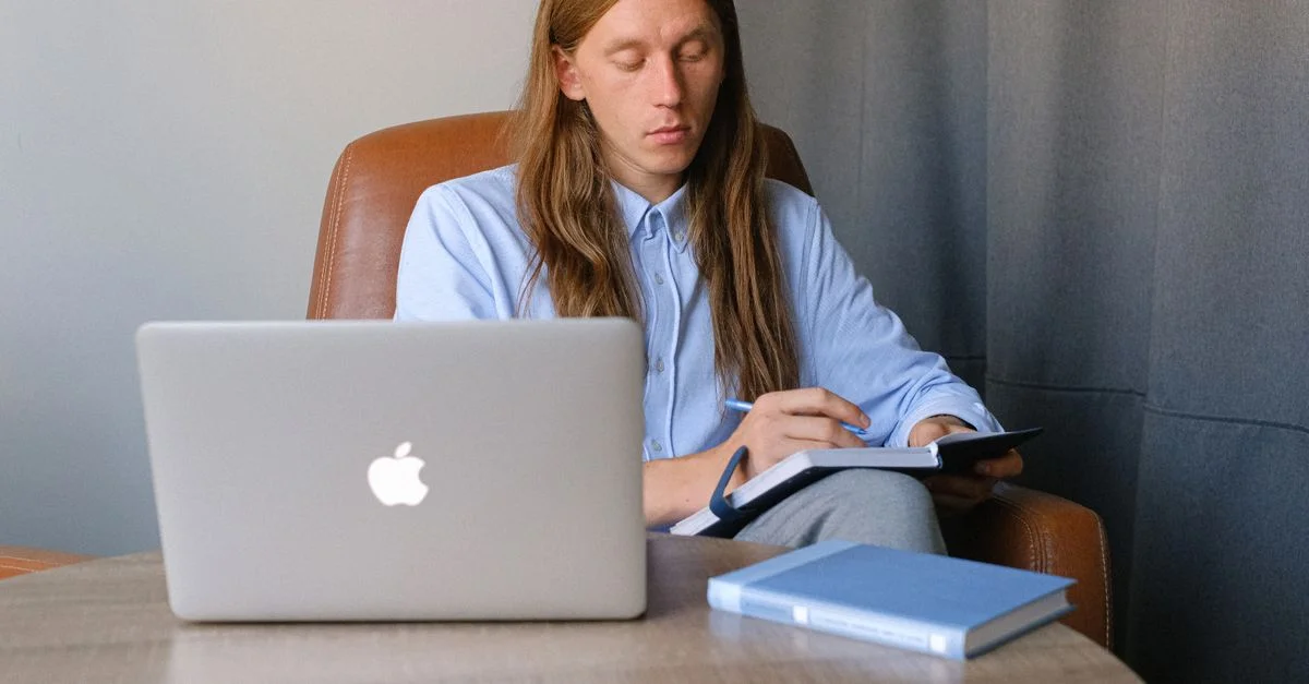 Jeune professionnel concentré prenant des notes à un bureau avec un ordinateur portable, illustrant la concentration et l'efficacité au travail.